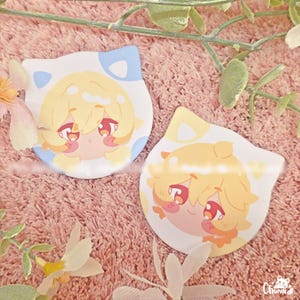 Può includere: Due adesivi a forma di gatto con illustrazioni in stile anime. Uno ha un orecchio blu e l'altro giallo. Entrambi presentano un personaggio biondo con occhi rossi e un'espressione carina. Su una superficie rosa strutturata.