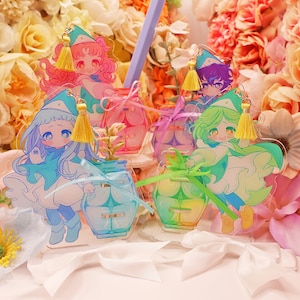 Puede incluir: Cuatro figuras de acrílico con personajes estilo anime con sombreros puntiagudos y capas, cada una con una base en forma de frasco. Las figuras tienen cabello y atuendos coloridos, acentuados con pequeñas borlas amarillas y cintas de acrílico.