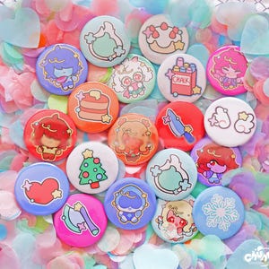 May include: A selection of colourful, round pin badges featuring cartoon illustrations. Designs include a Christmas tree, a heart, a snowflake, and characters with food and other objects. The badges are scattered on a bed of pastel confetti.