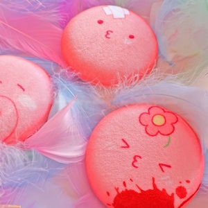 Pode incluir: Três itens redondos e rosa de pelúcia com rostos de desenhos animados e vários desenhos, incluindo uma bandagem, uma flor e um coração, repousam sobre uma cama de penas em tons pastel. Os itens têm detalhes bordados e são em um estilo caprichoso.