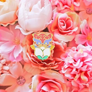 Puede incluir: Un colorido pin de esmalte con un personaje de dibujos animados con detalles en rojo, azul y amarillo, sobre un fondo de flores rosas y melocotón. El diseño del pin es detallado y artístico, ideal para añadir un toque personal a cualquier atuendo o accesorio.
