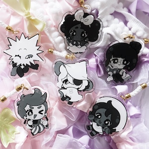 Puede incluir: Una colección de colgantes acrílicos con personajes de dibujos animados en negro, blanco y gris. Cada colgante tiene un diseño único y está unido a un cordón blanco con un cierre dorado. El fondo incluye cintas y lazos en rosa pastel, blanco y lavanda.