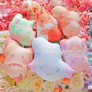 Puede incluir: Colección de seis peluches en varios colores pastel, incluyendo rosa, naranja, verde y azul. Cada peluche presenta un diseño único de personaje de dibujos animados con detalles bordados. Los juguetes están dispuestos sobre un lecho de flores coloridas.