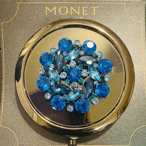 May include: A gold-toned compact mirror with a cluster of blue and teal gemstones. The compact is round and features the word "MONET" in gold lettering on the top of the box.