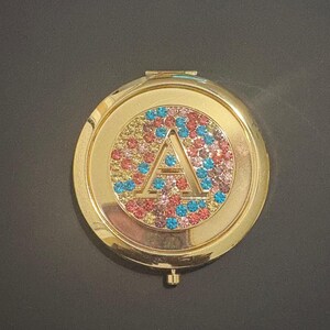 May include: A gold-colored, round compact mirror with a jeweled design. The center features a large letter "A" outlined in clear stones, surrounded by a circle of colorful rhinestones in shades of pink, blue, and yellow.