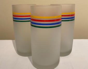 May include: Three frosted glass tumblers with horizontal bands of color: green, blue, red, and yellow. The glasses are cylindrical and have a simple, elegant design, suitable for serving beverages.