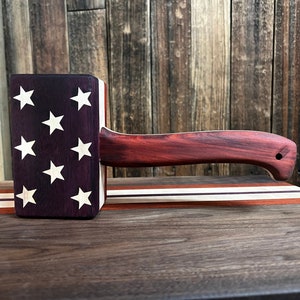May include: A wooden mallet with a red handle and a purple and white face with white stars. The mallet is sitting on a wooden surface.