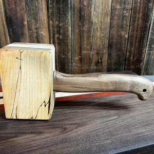 May include: A wooden mallet with a light brown handle and a square head made of light brown wood with dark brown streaks. The head has a white stripe across the top.