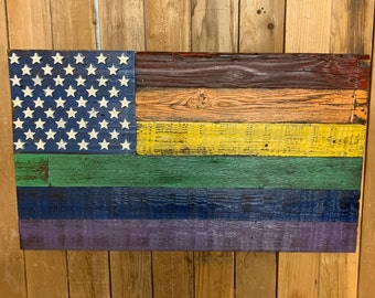 May include: A wooden American flag with a rainbow pride flag design. The flag is made of reclaimed wood and has a distressed look. The stars are white and the stripes are red, orange, yellow, green, blue, and purple.