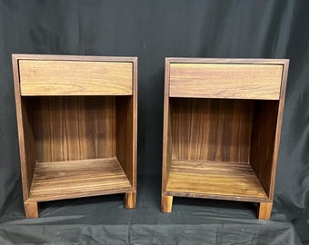 Puede incluir: Dos mesitas de noche de madera a juego con un acabado marrón cálido. Cada mesita de noche tiene un cajón en la parte superior y un estante abierto debajo. Las mesitas de noche están construidas con vetas de madera verticales y tienen un diseño simple y moderno.