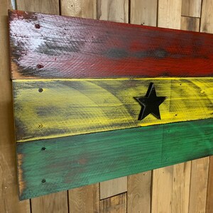 May include: A wooden sign painted in the colors of the Ghanaian flag. The sign is made of reclaimed wood and has a distressed look. The flag is red, yellow, and green with a black star in the center.