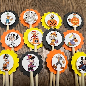 Puede incluir: Una colección de adornos para cupcakes con temática de Halloween con varios personajes de Disney disfrazados. Cada adorno tiene un borde festoneado en negro, naranja o amarillo, con una imagen circular de un personaje en un palito de madera.