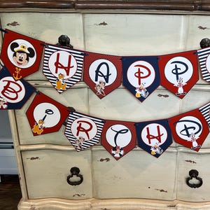 May include: A festive banner with red, white, and navy blue pennants. Each pennant features a letter spelling "HAPPY BIRTHDAY" and Disney characters like Mickey and Minnie Mouse. The banner is decorated with nautical elements.