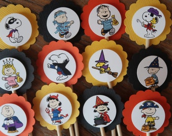May include: Twelve round cupcake toppers featuring Peanuts characters dressed in Halloween costumes. The toppers are on black, orange, and yellow scalloped circles. The characters include Snoopy, Charlie Brown, Lucy, and Linus.