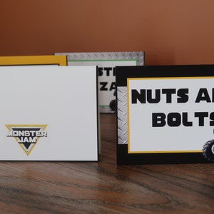 May include: Black and yellow sign with the text "NUTS AND BOLTS" in white. A red monster truck with black wheels is on the right side of the sign. The sign has a silver background with a diamond pattern.