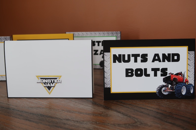 May include: Black and yellow sign with the text "NUTS AND BOLTS" in white. A red monster truck with black wheels is on the right side of the sign. The sign has a silver background with a diamond pattern.