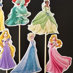 May include: Set of seven Disney princess cake toppers. The princesses are Snow White, Ariel, Tiana, Rapunzel, Elsa, Belle, and Cinderella. Each princess is wearing her signature dress and has a wooden stick attached to the back.