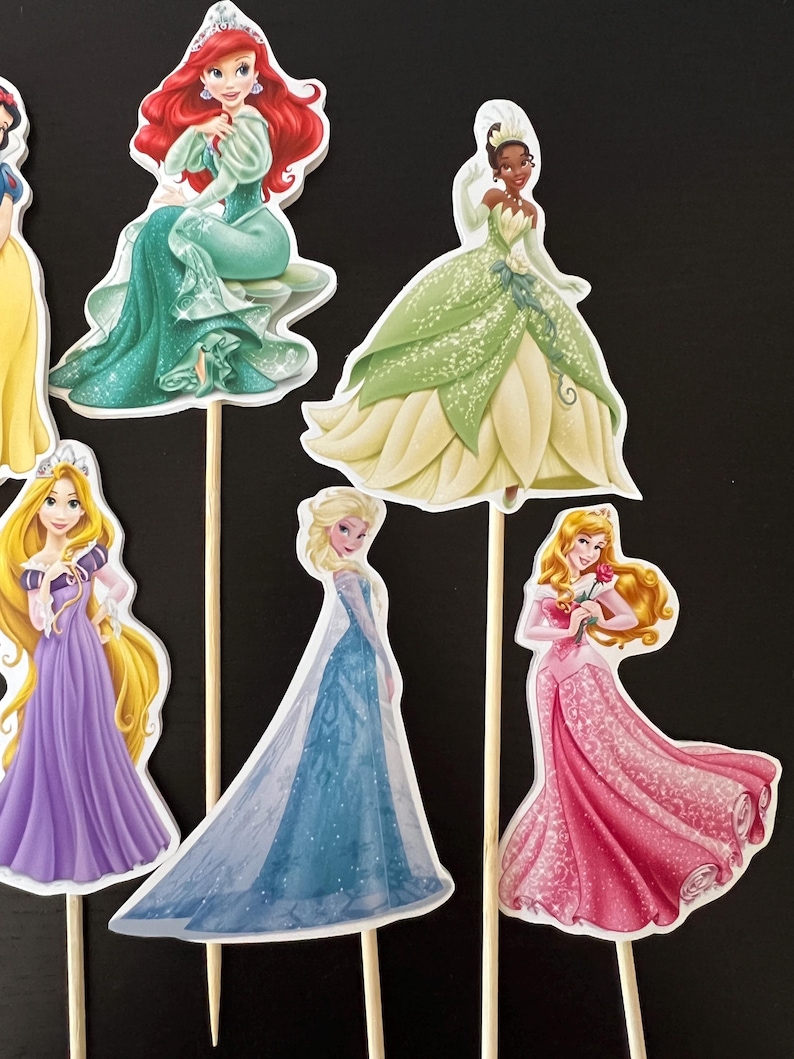 May include: Set of seven Disney princess cake toppers. The princesses are Snow White, Ariel, Tiana, Rapunzel, Elsa, Belle, and Cinderella. Each princess is wearing her signature dress and has a wooden stick attached to the back.