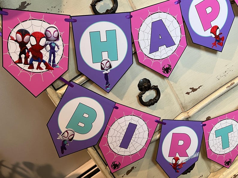 May include: A decorative party banner featuring pentagonal flags in shades of pink and purple. The flags display the letters H, A, P, B, I, R, T, and characters from Spidey and His Amazing Friends, including spider web patterns and small spider icons.