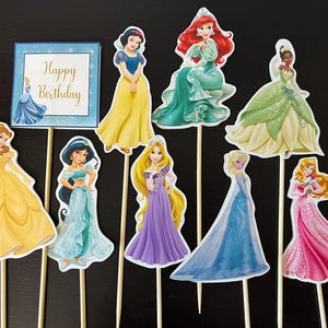 May include: Set of 9 Disney princess cake toppers featuring Cinderella, Belle, Ariel, Snow White, Jasmine, Rapunzel, Tiana, Elsa, and Aurora. The toppers are printed on cardstock and have wooden sticks attached. The toppers are perfect for a princess-themed birthday party.