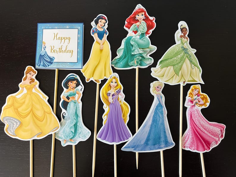 May include: Set of 9 Disney princess cake toppers featuring Cinderella, Belle, Ariel, Snow White, Jasmine, Rapunzel, Tiana, Elsa, and Aurora. The toppers are printed on cardstock and have wooden sticks attached. The toppers are perfect for a princess-themed birthday party.