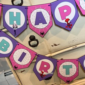 May include: A decorative party banner featuring purple and pink pennants with spiderweb patterns and cartoon superhero characters. Each pennant displays a letter in teal or pink, spelling out HAPPY BIRTH, with small black spider icons.