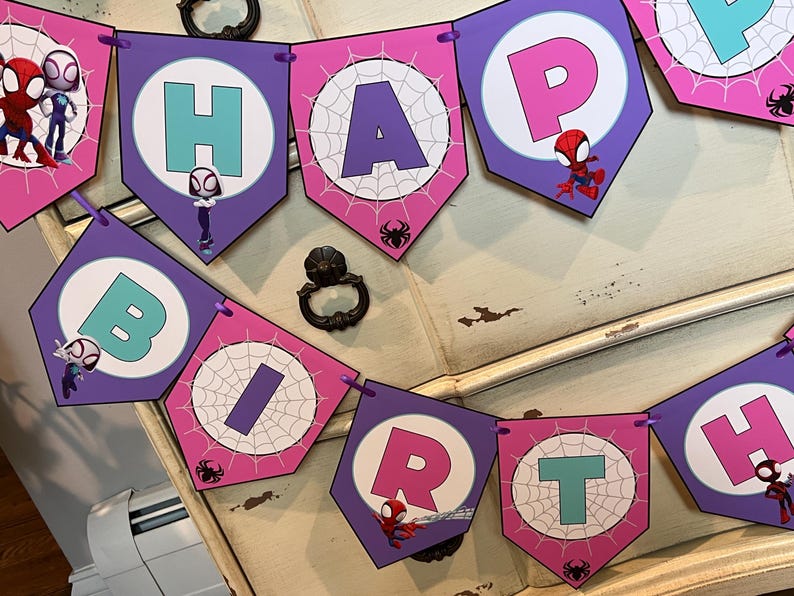 May include: A decorative party banner featuring purple and pink pennants with spiderweb patterns and cartoon superhero characters. Each pennant displays a letter in teal or pink, spelling out HAPPY BIRTH, with small black spider icons.