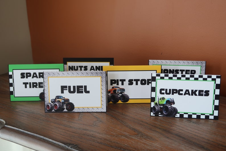 May include: Five monster truck themed food labels for a party. The labels are printed on cardstock and feature a monster truck graphic. The labels read "Spare Tire", "Fuel", "Pit Stop", "Monster Cupcakes", and "Nuts and Bolts".