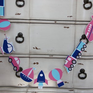May include: A garland of transportation-themed cutouts in pink, blue, and white. The garland includes a car, boat, rocket, hot air balloon, airplane, train, and helicopter. The garland is strung on a red string and is displayed against a white dresser.