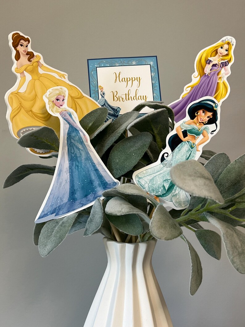May include: A bouquet of artificial greenery in a white vase with four princess character toppers. The toppers feature Belle, Elsa, Rapunzel, and Jasmine. The card says "Happy Birthday".