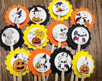 May include: Twelve Halloween-themed cupcake toppers featuring Snoopy dressed in various costumes, including a witch, a vampire, a mummy, and a pirate. Each topper is on a wooden stick and has a scalloped edge.