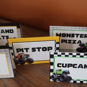 May include: Five monster truck themed food signs with black and white checkered borders. The signs read "Nuts and Bolts", "Pit Stop", "Monster Pizza", "Cupcakes", and "Fuel".