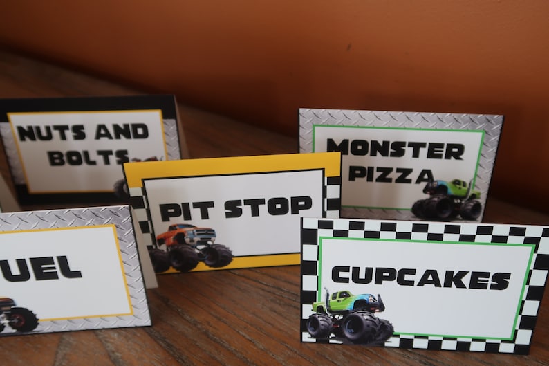 May include: Five monster truck themed food signs with black and white checkered borders. The signs read "Nuts and Bolts", "Pit Stop", "Monster Pizza", "Cupcakes", and "Fuel".