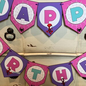 May include: A decorative birthday banner featuring pentagonal flags in shades of purple and pink. Each flag displays a letter spelling out HAPPY BIRTHDAY, accented with white spiderweb patterns, small spider icons, and cartoon superhero characters.