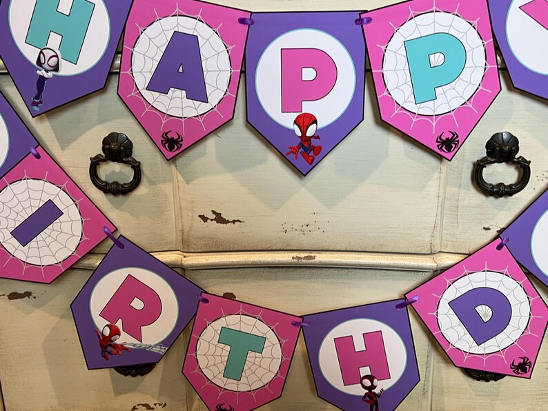 May include: A decorative birthday banner featuring pentagonal flags in shades of purple and pink. Each flag displays a letter spelling out HAPPY BIRTHDAY, accented with white spiderweb patterns, small spider icons, and cartoon superhero characters.