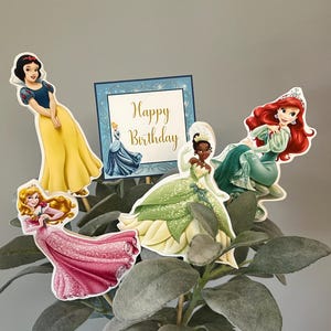 May include: A set of five Disney princess cake toppers featuring Snow White, Cinderella, Tiana, Ariel, and a sign that says "Happy Birthday".
