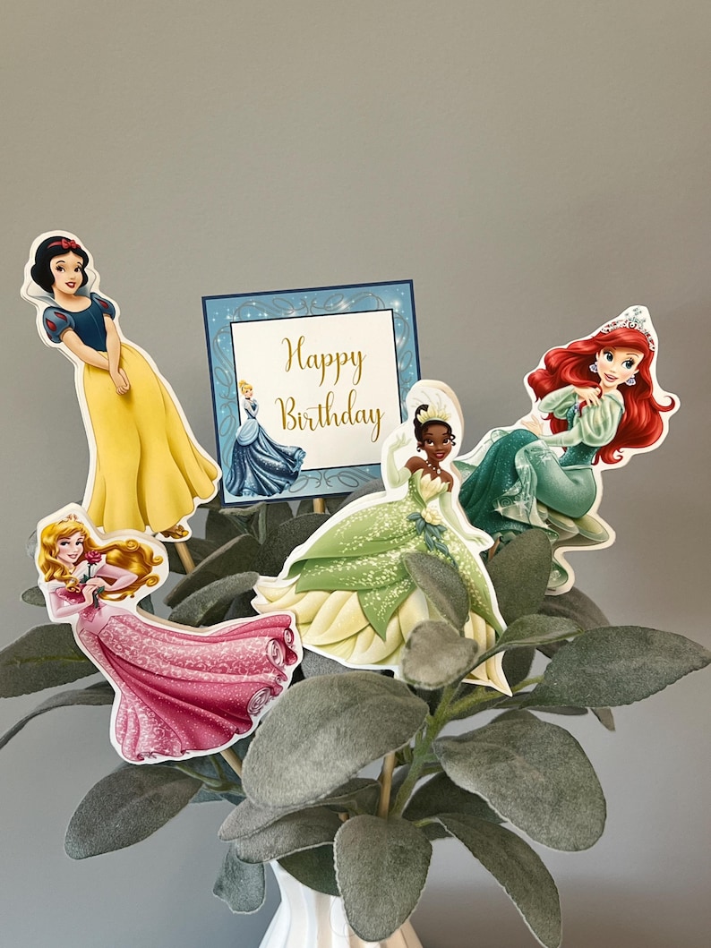 May include: A set of five Disney princess cake toppers featuring Snow White, Cinderella, Tiana, Ariel, and a sign that says "Happy Birthday".