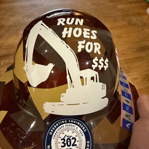 May include: A brown and tan camouflage hard hat featuring a white graphic of an excavator and the text RUN HOES FOR $$$. A circular sticker at the bottom displays the text OPERATING ENGINEERS CHARTERED AUGUST 6, 1905, 302, LABOR OMNIA VINCIT.