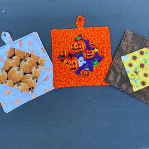 May include: Three square fabric pot holders with hanging loops. The left one is white with orange leaves and a pumpkin center. The middle one is orange with a purple center featuring jack-o'-lanterns and ghosts. The right one is brown with sunflowers.