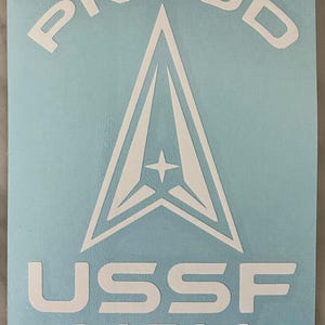 May include: White decal on a light blue background. The decal features the word "PROUD" arched across the top, above a stylized white emblem with a star. Below the emblem, the text reads "USSF MOM".