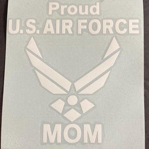 May include: White sticker with the text "Proud U.S. AIR FORCE MOM" and a stylized emblem. The emblem features a central star shape with upward-pointing wings. The sticker is on a light gray background.