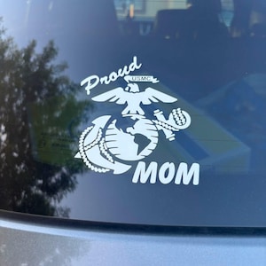 May include: A white vinyl decal applied to a car window featuring the United States Marine Corps Eagle, Globe, and Anchor emblem. The text Proud is arched above the emblem, and the word MOM is printed in large white capital letters below it.