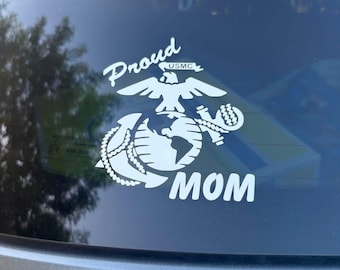 May include: A white vinyl decal applied to a vehicle window featuring the United States Marine Corps emblem, which includes an eagle, globe, and anchor. The text Proud is arched above the emblem, and the word MOM is positioned below it.