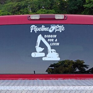 May include: A white vinyl decal applied to a red truck rear window. The decal features a graphic of an excavator lifting a pipe, with the text Pipeline Life above and the words DIGGIN FOR A LIVIN positioned to the right of the excavator arm.