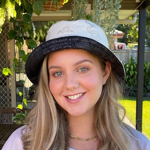 May include: A woman with long blonde hair is wearing a white bucket hat with a black snakeskin pattern around the brim. The hat has a black snake design on the front. She is wearing a white shirt with a gold chain necklace.