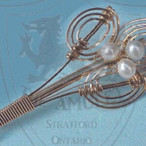 Peut inclure: Une broche en fil d'or avec trois perles blanches au centre. Le fil est torsadé en un motif décoratif ressemblant à une fleur.