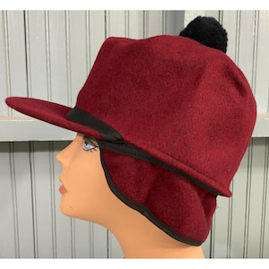 May include: A burgundy felt hat with a black pom-pom on top and a black ribbon around the brim. The hat has a wide brim and a rounded crown.