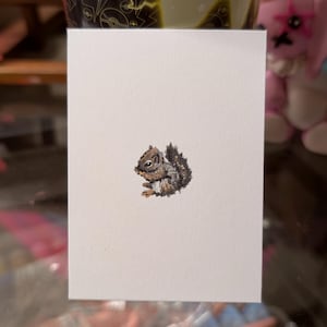 May include: A watercolor painting of a small squirrel on a white card. The squirrel is depicted in shades of brown, gray, and black, with a fluffy tail and a small nut in its paws. The artwork is detailed and realistic.