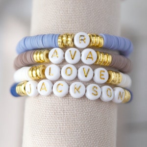 May include: A stack of colorful beaded bracelets with gold beads and white letter beads spelling out the names "Ava", "Love", and "Jackson".