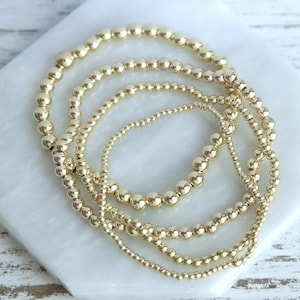 May include: Four gold beaded bracelets of varying sizes are arranged on a white marble surface. The bracelets feature small, round beads in a shiny gold color. The bracelets are stacked together, creating a layered look.